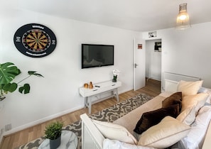 A living room with a dartboard, a flat-screen TV, a white sofa, a small table with decorative items, and a potted plant. - A lovely cosy property in a lovely location with stunning scenery. (Kenley, near Surrey)