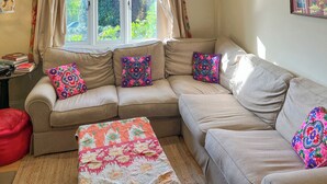 Un salon chaleureux avec un canapé beige, des coussins colorés, une petite table avec des livres et une fenêtre avec des rideaux.