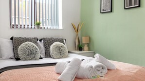 Un lit bien fait, avec des serviettes blanches, une couverture rose à motifs et des coussins à motifs. On y trouve une petite table de chevet avec une lampe et un vase contenant des plantes séchées. Les murs de la pièce sont d’un vert clair et il y a une fenêtre avec des stores verticaux.