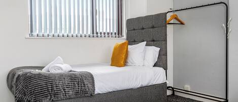 A neatly made bed with a grey headboard, white bedding, and a yellow pillow. A grey upholstered bench is against the wall, and a clothing rack with a yellow garment is visible.