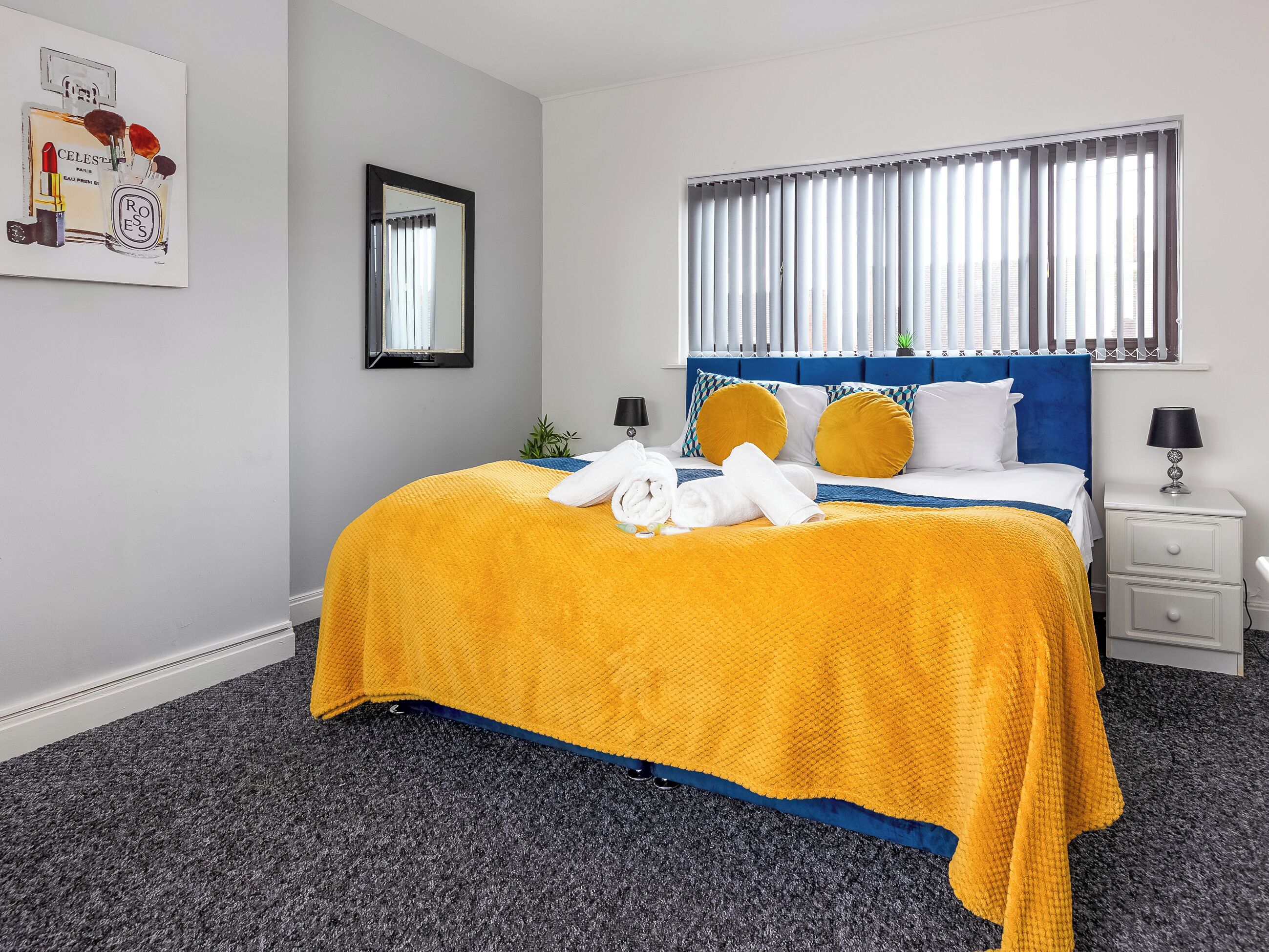 Een hotelkamer met een bed, nachtkastje, lamp, spiegel en raam met jaloezieën.
