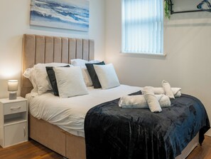 A neatly made bed with a dark blue bedspread, white pillows, and a headboard. There is a bedside table with a lamp and a small plant on the window sill.