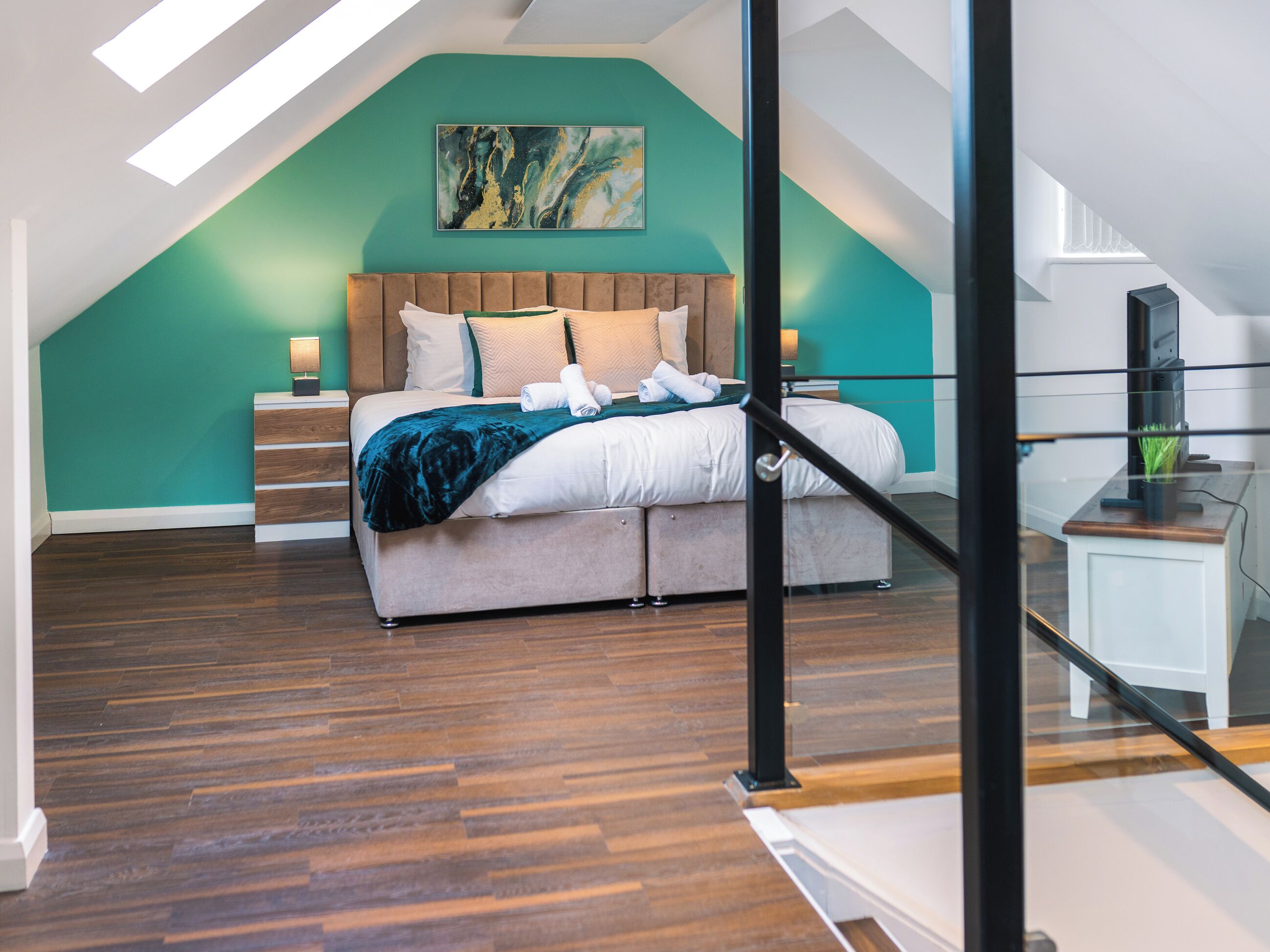A bedroom with a sloped ceiling, a bed with a wooden headboard, a teal accent wall, and a painting above the bed.
