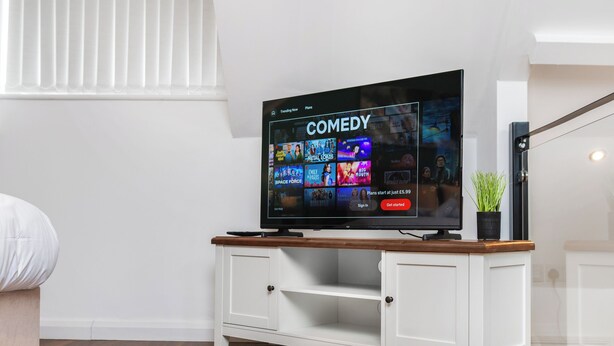 A flat-screen TV on a white entertainment unit with a wooden top, displaying a comedy channel interface.