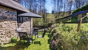 A stone building with a patio area featuring a table and chairs, surrounded by a wooden fence and lush greenery.