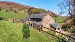 A stone farmhouse with a sloped roof, surrounded by a wooden fence, set in a rural landscape with green fields and trees.