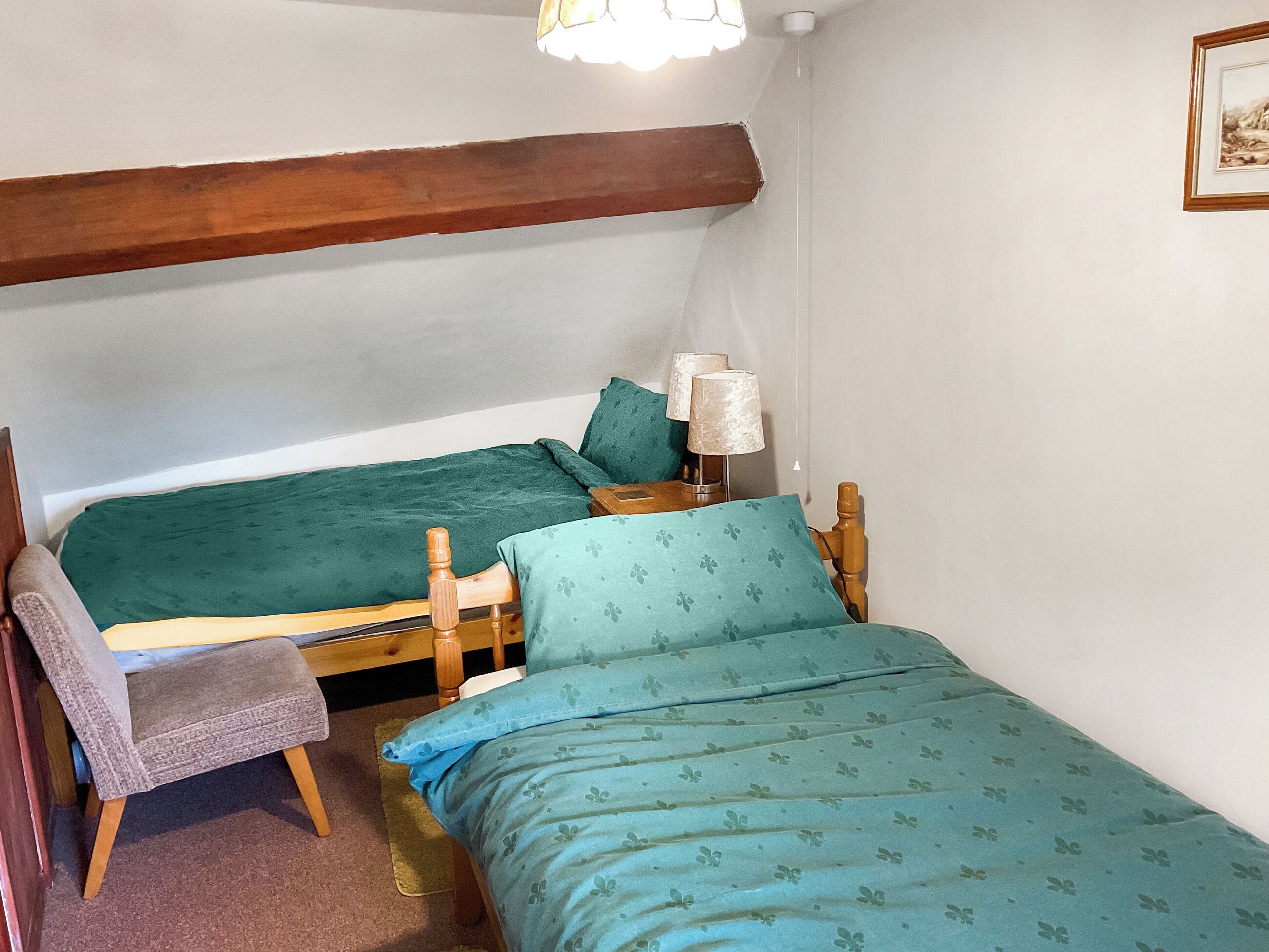 A small, cozy room with two single beds, a wooden chair, a nightstand with a lamp, and a framed picture on the wall.