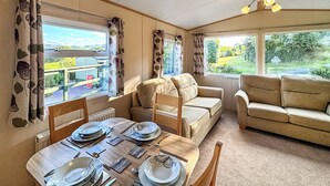 A cozy living room with a dining area, featuring a sofa set, a dining table set for four, and large windows offering a view of the outdoors. - 2 bedroom accommodation in Machynlleth (Machynlleth)