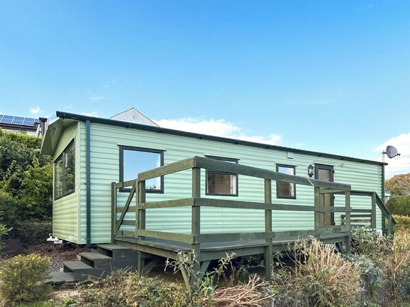 A small, light green mobile home with a wooden deck and a satellite dish.