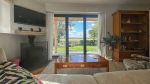 A living room with a TV, a wooden coffee table, a sofa, a bookshelf, and a large window with a view of greenery. - 4 bedroom accommodation in Royal Wootton Bassett (Royal Wootton Bassett)