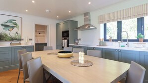 A modern kitchen with a dining area, featuring a wooden table, grey chairs, and a large window with a view of greenery. - 4 bedroom accommodation in Royal Wootton Bassett (Royal Wootton Bassett)