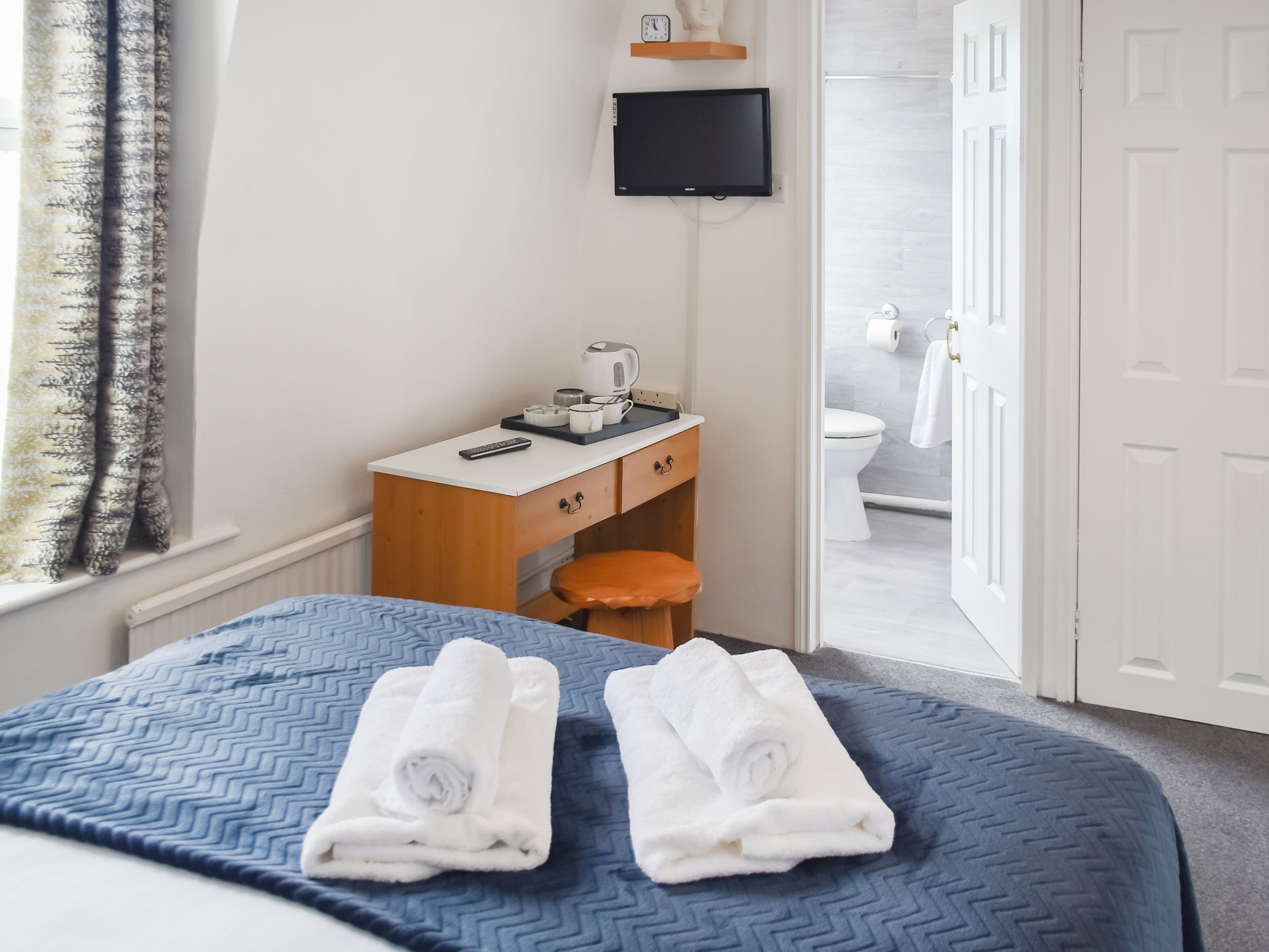 A hotel room with a bed, a desk with a kettle, a TV, and a bathroom visible through an open door.
