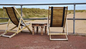 Zwei Holzliegen mit Metallstreben, ein kleiner Holztisch und ein Metallgeländer mit Blick auf einen Sandstrand.
