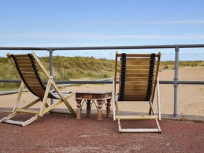 Zwei Holzliegen mit Metallstreben, ein kleiner Holztisch und ein Metallgeländer mit Blick auf einen Sandstrand.