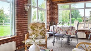 Une pièce avec un mur en briques, de grandes fenêtres, un fauteuil en osier, une table recouverte d’une nappe à motifs floraux et un vase rempli de fleurs.