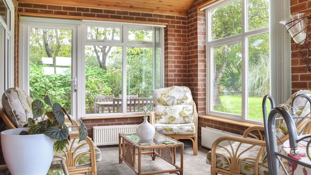 Une véranda meublée de mobilier en osier, dotée de grandes fenêtres et offrant une vue sur un jardin.