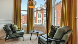A room with two green armchairs, a small table, and a floor lamp. Large windows with curtains offer a view of red brick buildings.