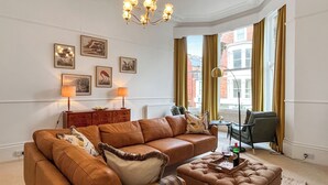 A modern living room with a brown leather sofa, a matching ottoman, and a dining table with a tray. There are framed pictures on the wall, a chandelier, and a view of the city through large windows.