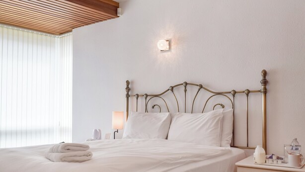 Ein ordentlich bezogenes Bett mit weißen Leinen, ein Nachttisch mit Lampe und eine Wandleuchte.