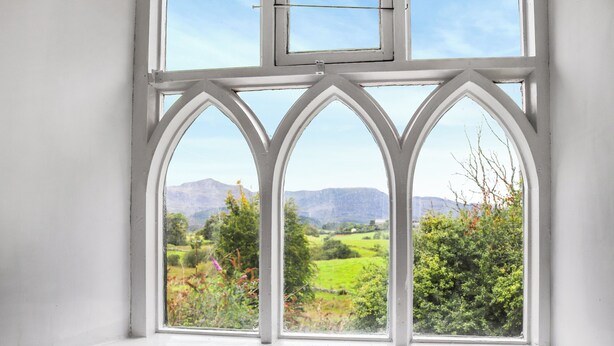 Una ventana con marco blanco y tres arcos, con vistas a un paisaje verde con montañas.