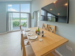 A dining area with a wooden table set for four, a large window with a view of greenery, and a flat-screen TV mounted on the wall.