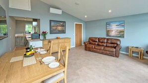 A modern living room with a brown leather sofa, a dining table with chairs, and a kitchen area in the background. - Grey Rocks Apartment (Kippford)