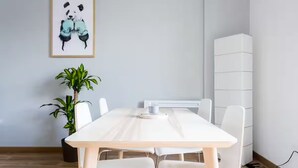 A minimalist room with a white table, white chairs, a potted plant, and a framed artwork on the wall.