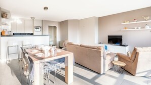 Un salon moderne avec un canapé beige, une table à manger avec des chaises et un coin cuisine doté de meubles blancs et d’un téléviseur.