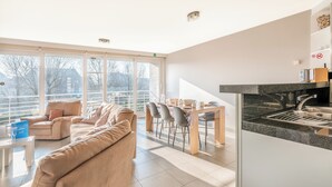 Un salon moderne avec un canapé beige, un coin repas doté d’une table en bois et une cuisine avec un plan de travail en granit.