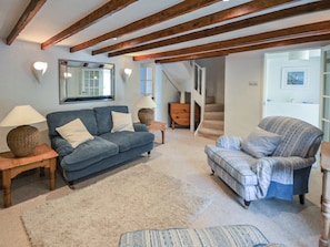 A living room with a blue sofa, a patterned armchair, wooden beams on the ceiling, a mirror, and a staircase leading to another room.