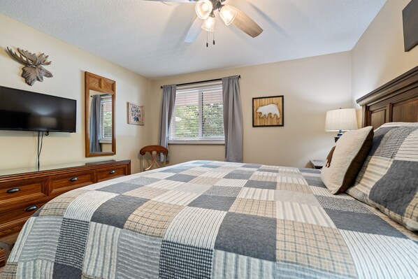 A bedroom with a large bed, a ceiling fan, a TV mounted on the wall, and a window with curtains.
