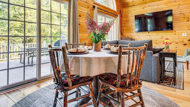 Interior de una cabaña de madera con una mesa de comedor dispuesta para cuatro personas, rodeada de sillas de madera.