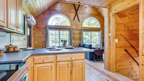 Interior de una cabaña de madera con cocina, que incluye armarios de madera, una encimera y una estufa. Dos ventanas arqueadas ofrecen luz natural y vistas al exterior.
