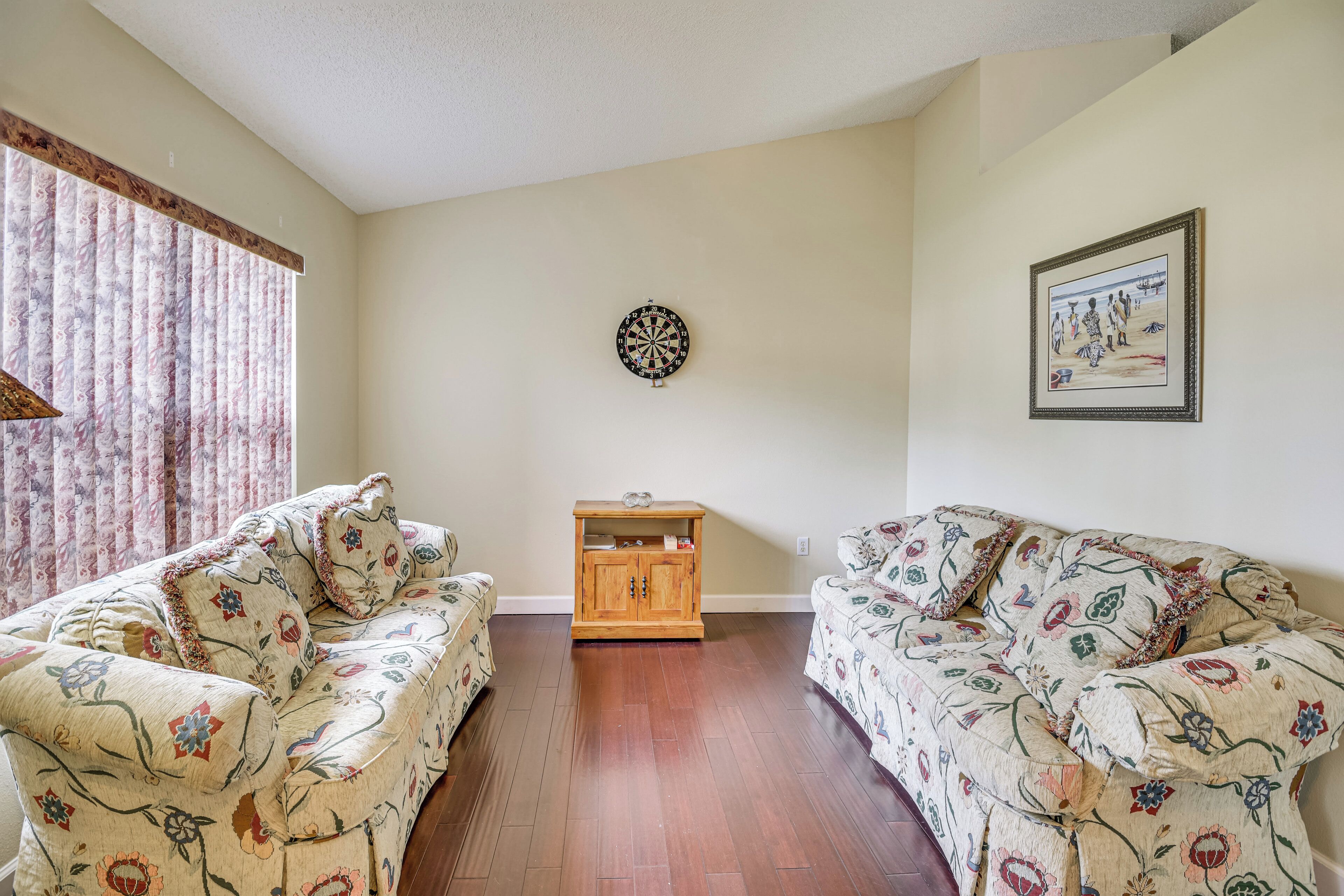 A living room with two floral-patterned sofas, a wooden cabinet, a dartboard, and a framed picture on the wall.
