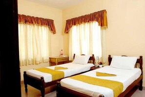 Habitación de hotel con dos camas individuales, cada una con estructura de madera y ropa de cama blanca. Hay dos mesitas de noche con lámparas, una pequeña mesa y una silla. Las paredes son color beige y las cortinas tienen un ribete marrón.