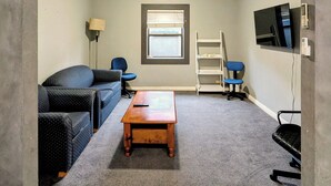 Ein Zimmer mit Deckenventilator, Jalousien am Fenster, einem an der Wand montierten Fernseher, einem Schreibtisch, einem Stuhl und einer Couch.