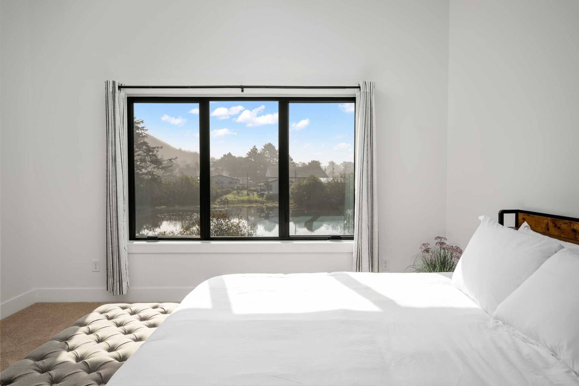 A bedroom with a large window, a bed, and a view of a lake and buildings.