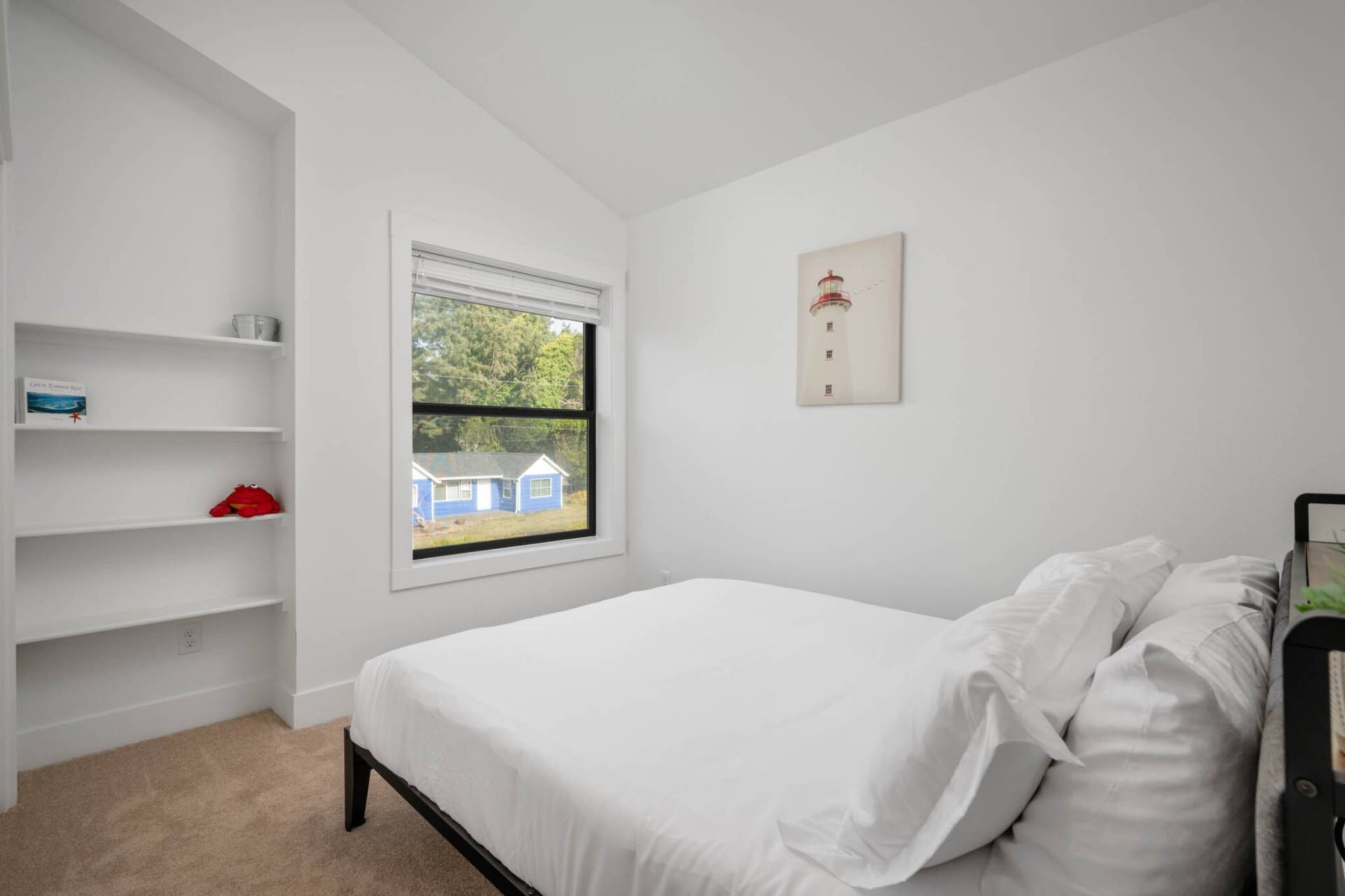 A bedroom with a single bed, a window with a view of a house, white walls, and a shelf with items.