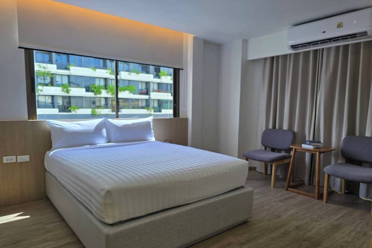 A modern hotel room with a large bed, two chairs, a small table, and a view of buildings through the window.