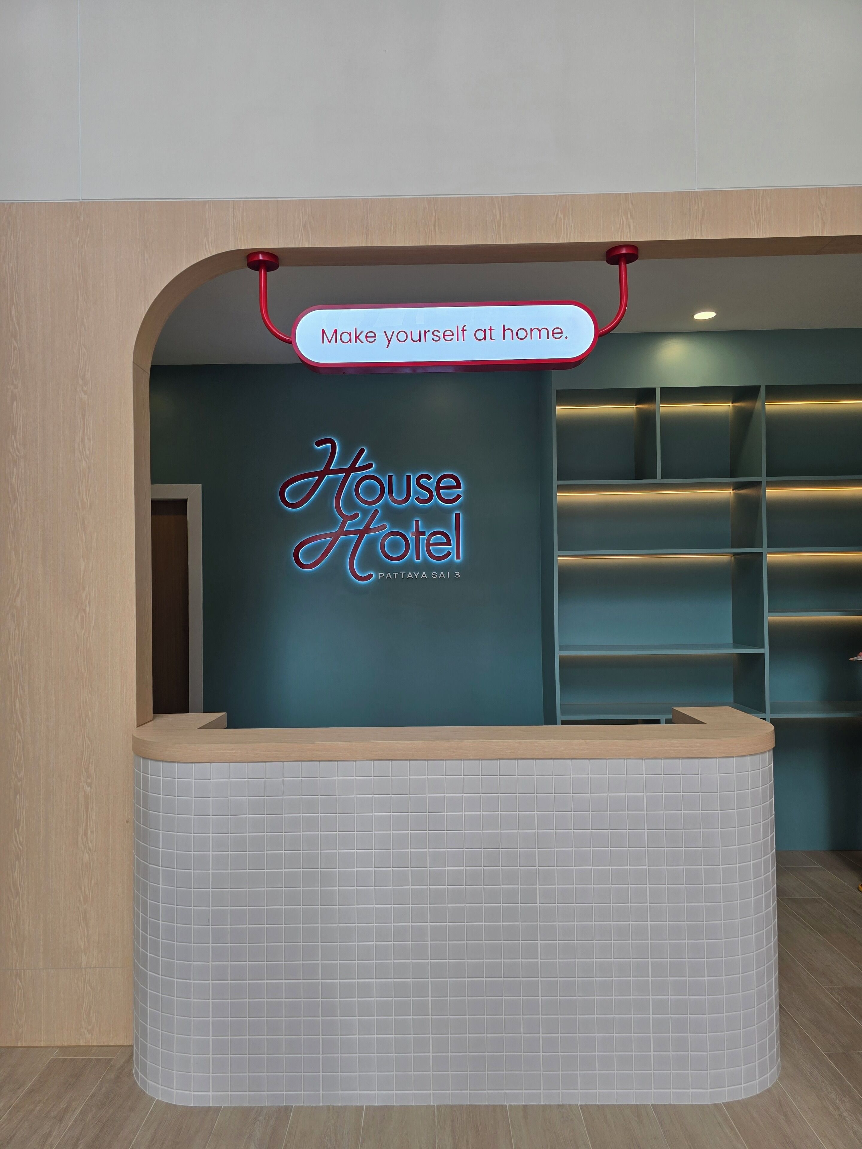 A reception desk with a neon sign reading 'House Hotel Pattaya South' and a sign above stating 'Make yourself at home.'