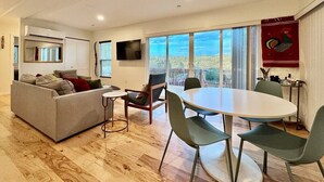 A modern living room with a sofa, a dining table with chairs, a TV mounted on the wall, and large windows offering a view of the outdoors. - Unique Container Home · 20 Acres · Starry Nights · EV Charger (Placerville)
