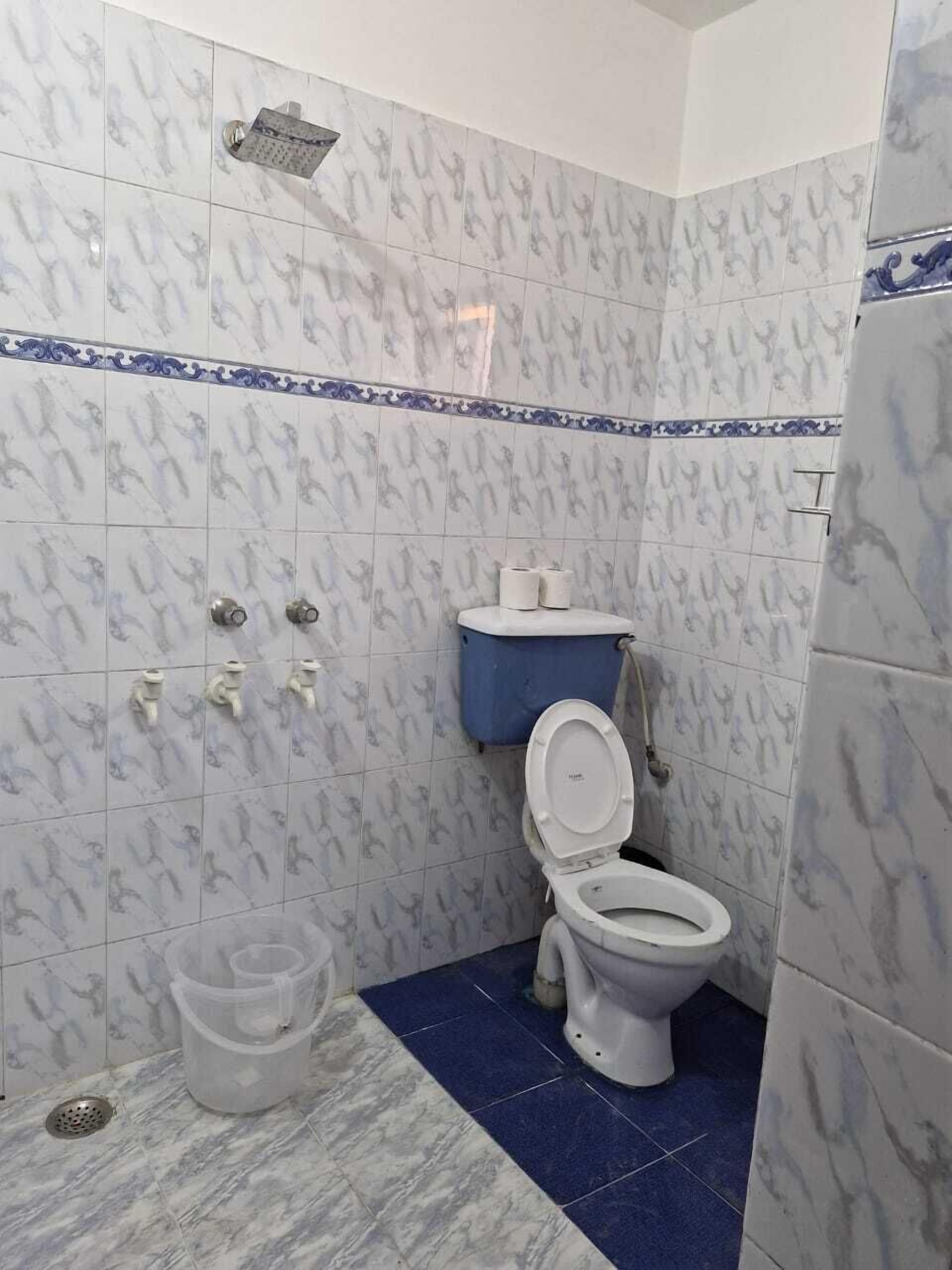 A bathroom with white marble tiles, a blue-tiled floor, a toilet, a shower, and a trash bin.