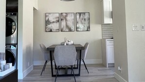 A modern kitchen with a built-in washer and dryer, a dining area with a gray table and chairs, and a hallway leading to a bathroom.
