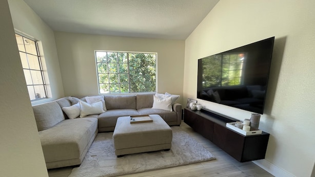 A modern living room with a sectional sofa, a flat-screen TV, and a large window with a view of trees.