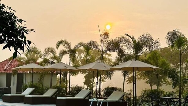 A swimming pool with lounge chairs and umbrellas, surrounded by palm trees and a building with a red roof. - Garden Centre Resort (Kaliganj)