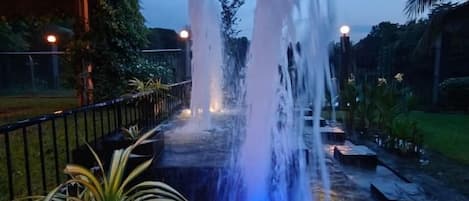 Una fontana esterna illuminata di notte, con più zampilli, circondata da vegetazione e un vialetto lastricato.