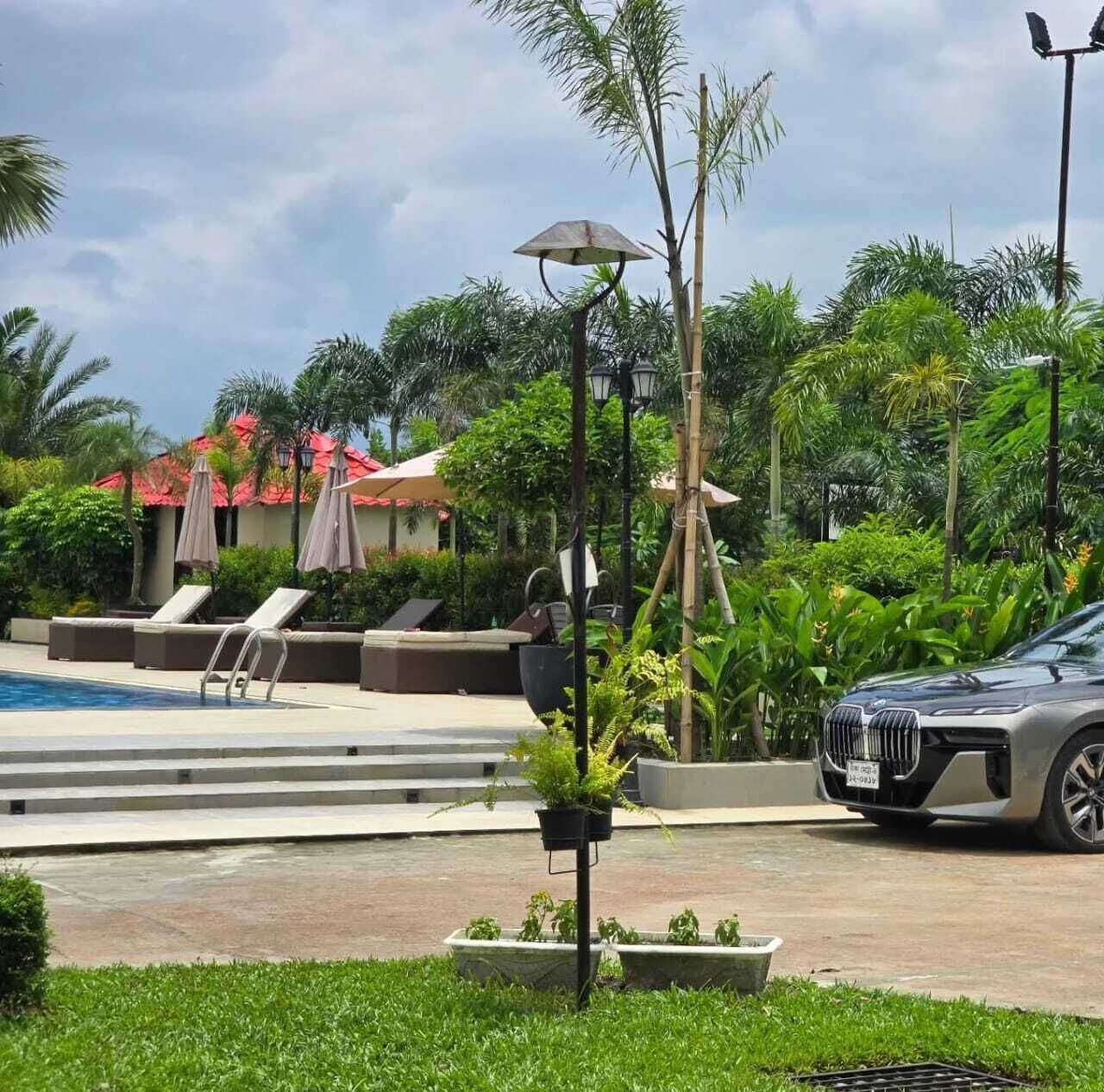 A pool area with lounge chairs and umbrellas, a car parked nearby, and lush greenery.