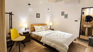 A bedroom with a wooden ceiling, a bed with floral bedding, a yellow chair, bedside lamps, and a small table. - Garden Centre Resort (Kaliganj)