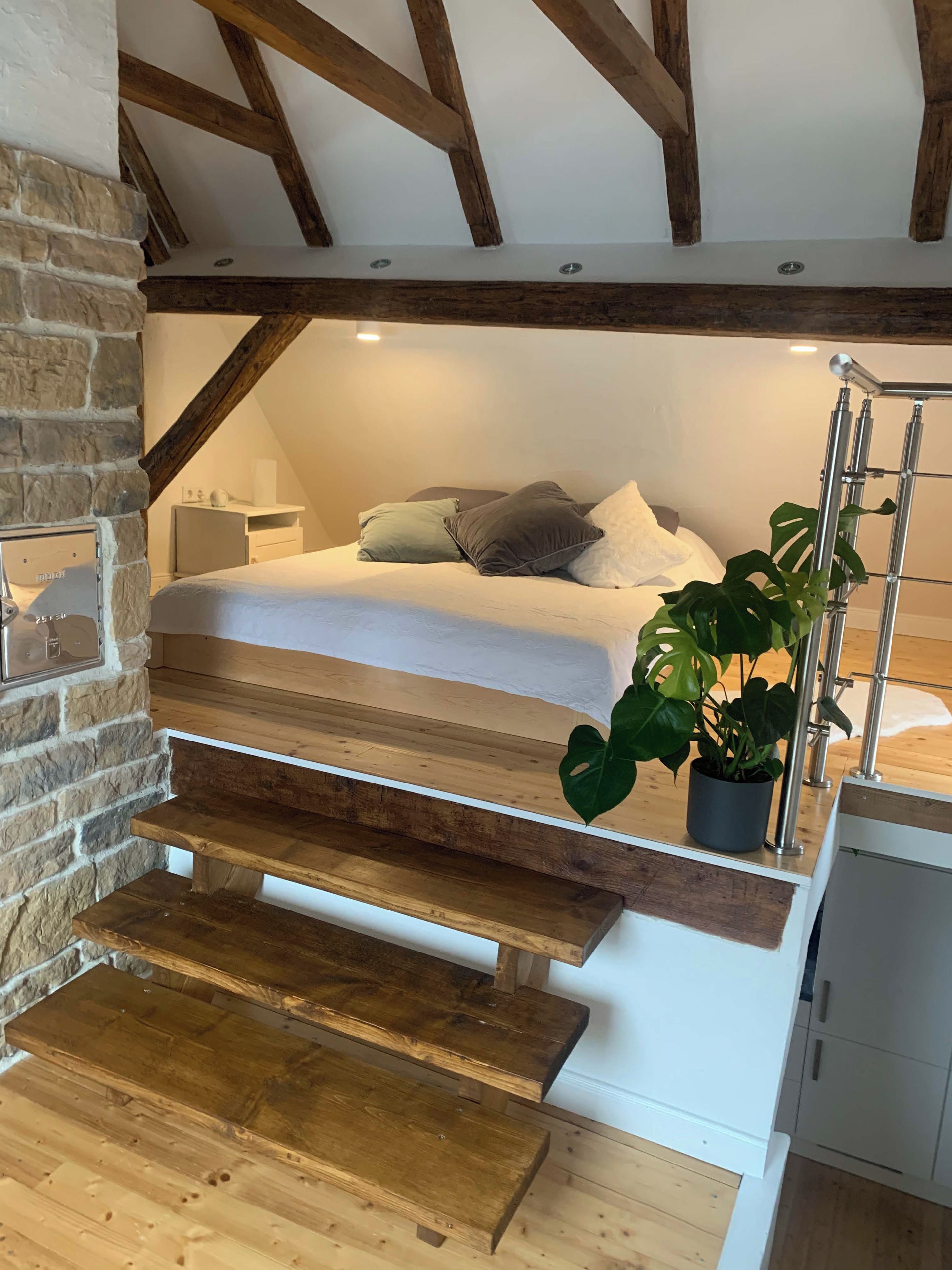 Une chambre à coucher avec un lit, des oreillers, une table de chevet, une plante en pot et un escalier menant à l’étage supérieur.