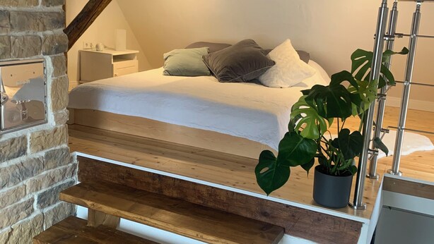 Ein Schlafzimmer mit Bett, Kissen, einem Nachttisch, einer Topfpflanze und einer Treppe, die in eine höhere Ebene führt.